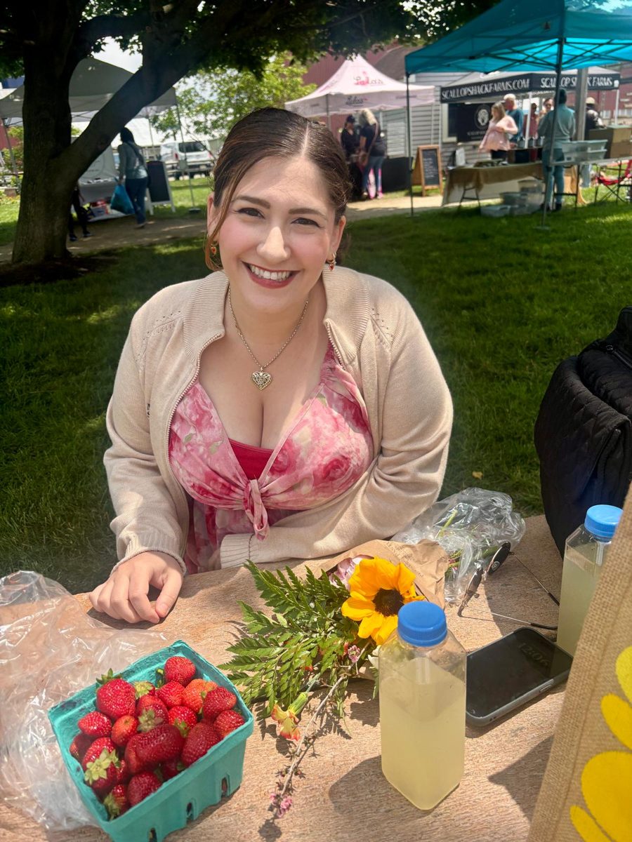 Val smiling at a picnic table at a farmers market, with strawberries, sunflowers, and lemonade