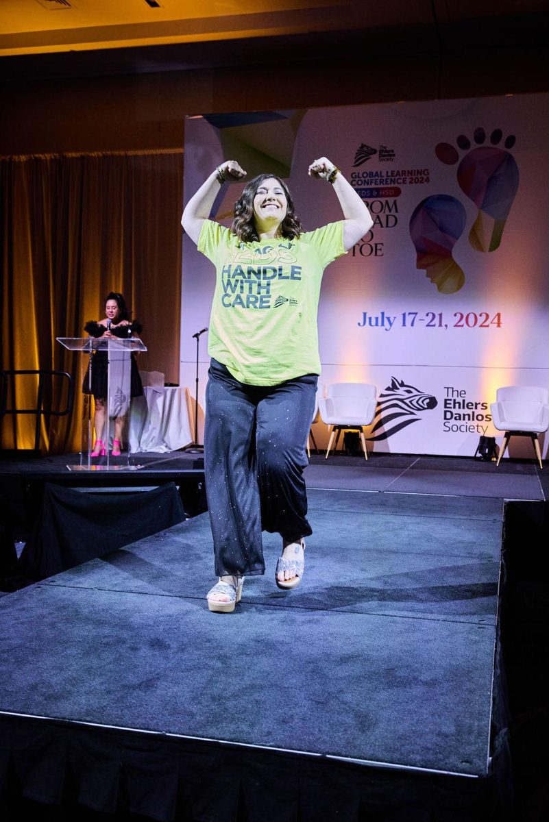 Val smiling and flexing on stage in a Handle With Care shirt at the 2024 Global Learning Conference